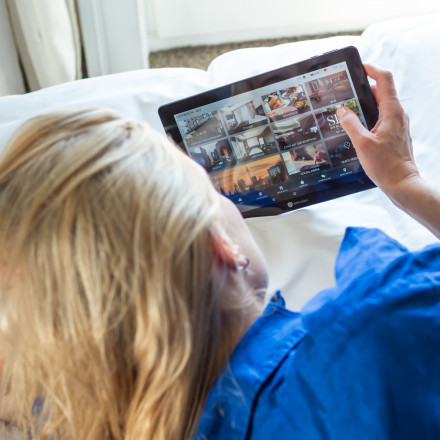 A person with blonde hair lies on a bed in a blue shirt, holding a tablet displaying multiple streaming videos.