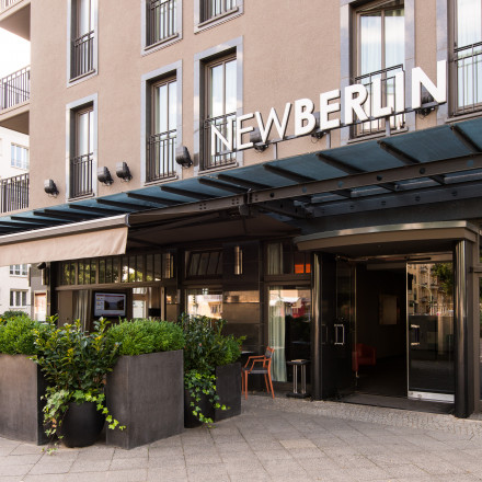 Straßenansicht eines modernen Hotels mit dem Schild „NEWBERLIN“, Pflanzen im Außenbereich und einem überdachten Eingang an der Hausnummer 24.