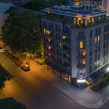 A modern hotel building on a city corner is lit up at night, with warm lights glowing from windows and streetlights illuminating the roads.