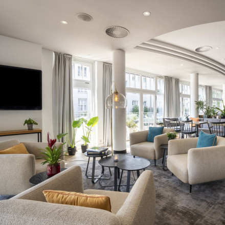 Modern lounge with beige armchairs, colorful pillows, potted plants, large windows, and a blue accent wall in the background.