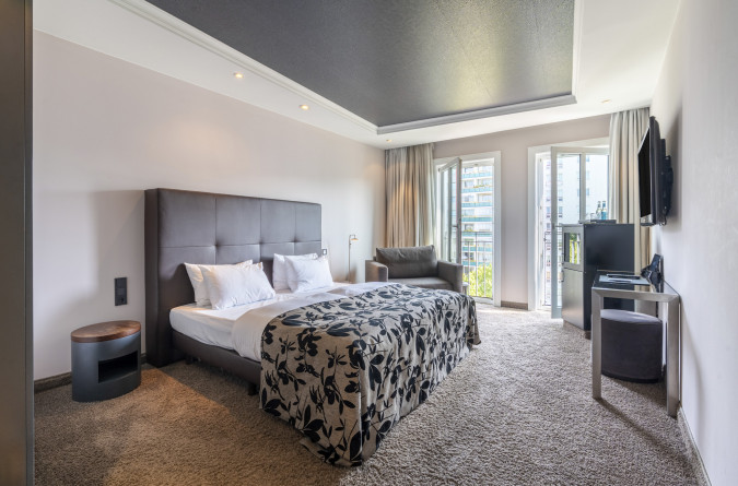 Modernes Hotelzimmer mit großem Bett, Schreibtisch, Fernseher, Sesseln und sanfter Beleuchtung, in neutralen und warmen Tönen gehalten.