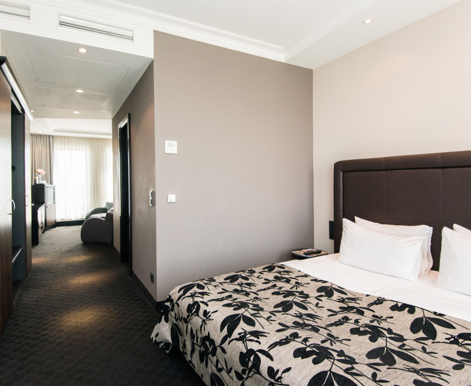 Modern hotel room with a large bed, a single bed, desk, TV, and balcony doors letting in natural light.